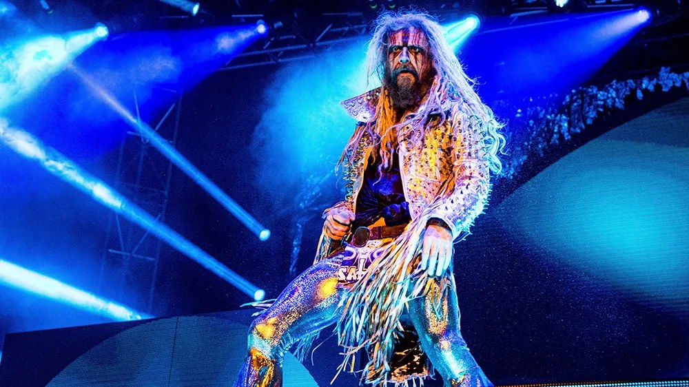 Performer with long hair and face paint in a sparkly spiked jacket on a stage lit by blue beams